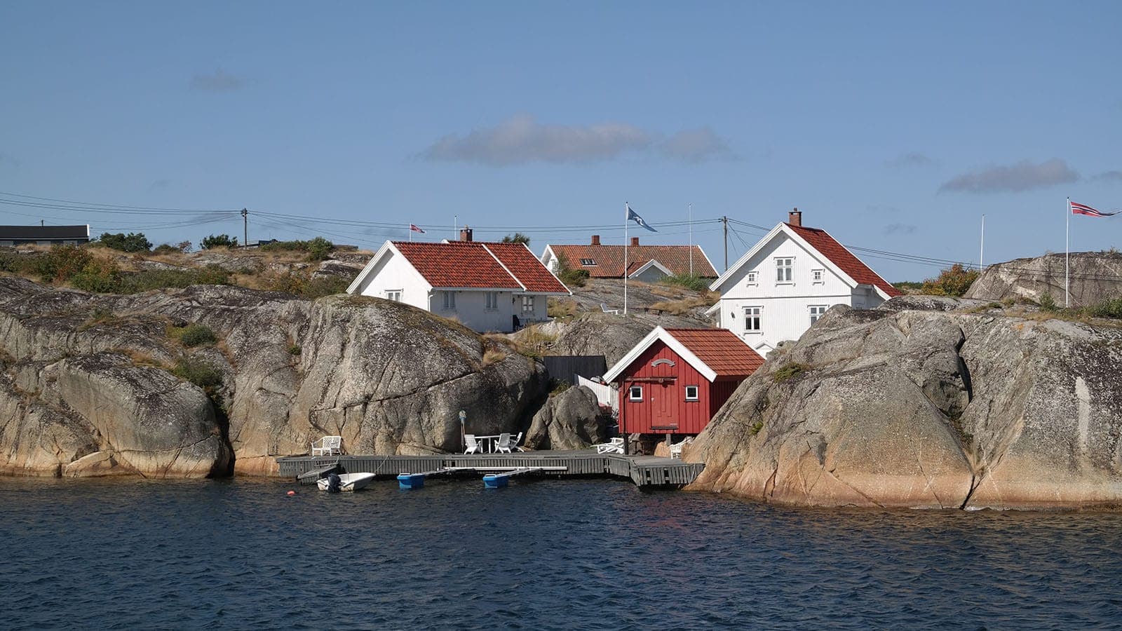 Hvitmalte hus og rød sjøbod mellom svabergene på Lauer, sett fra sjøen, med brygge og småbåter i forgrunnen.