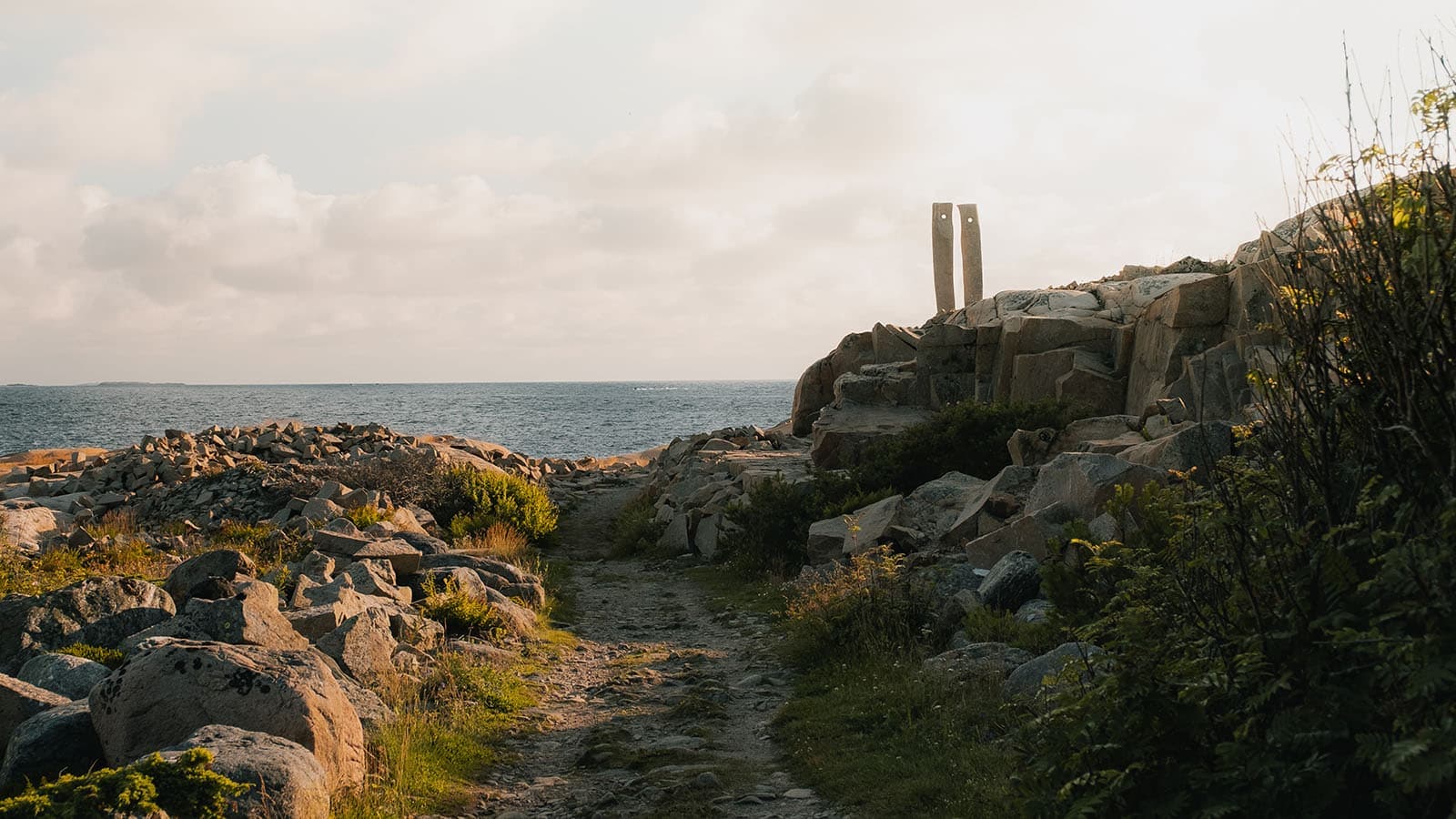 Kyststi som leder mot havet, med to høye stenskulpturer plassert på svaberget