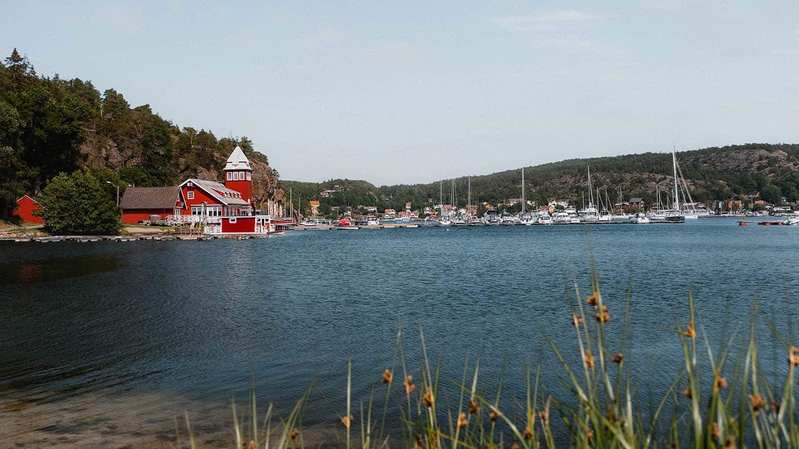 Rød bygning med tårn ved sjøen, med båthavn og bebyggelse i bakgrunnen på Hankø.