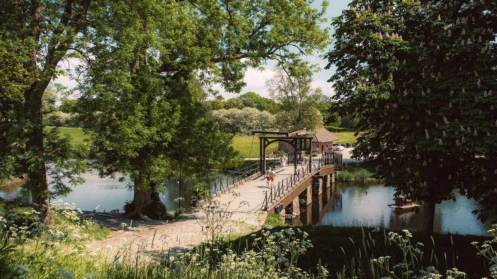 Folk som krysser Vindebrua inn til Gamlebyen i Fredrikstad på en solrik dag, omgitt av grønt landskap og vann.